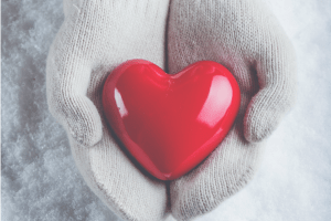 Female hands in white knitted mittens with a glossy red heart on a snow winter background.
