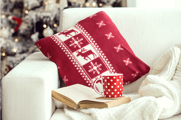 Opened book and a cup of tee on the cozy armchair with warm blanket and cushion on it near Christmas tree.