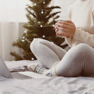 woman sitting on bed with hot drink and laptop, christmas tree in background