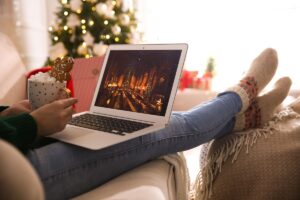 person with laptop in lap with christmas movie on it, christmas tree in the background, and they are holding a cup of hot chocolate