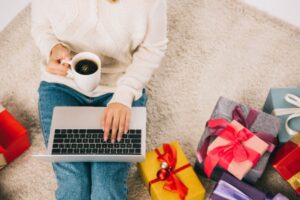 woman holding coffee with gifts around here while on laptop