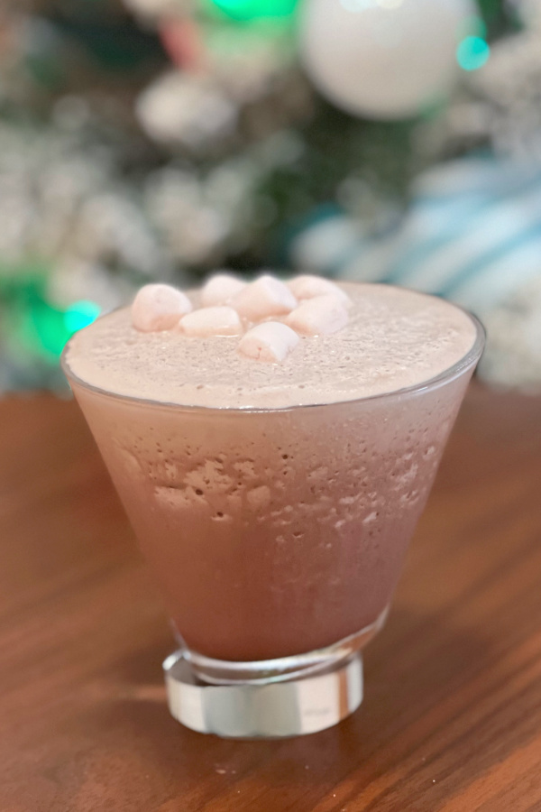 frozen hot chocolate martini in martini glass with mini marshmallows on top in front of flocked christmas tree blurred in background