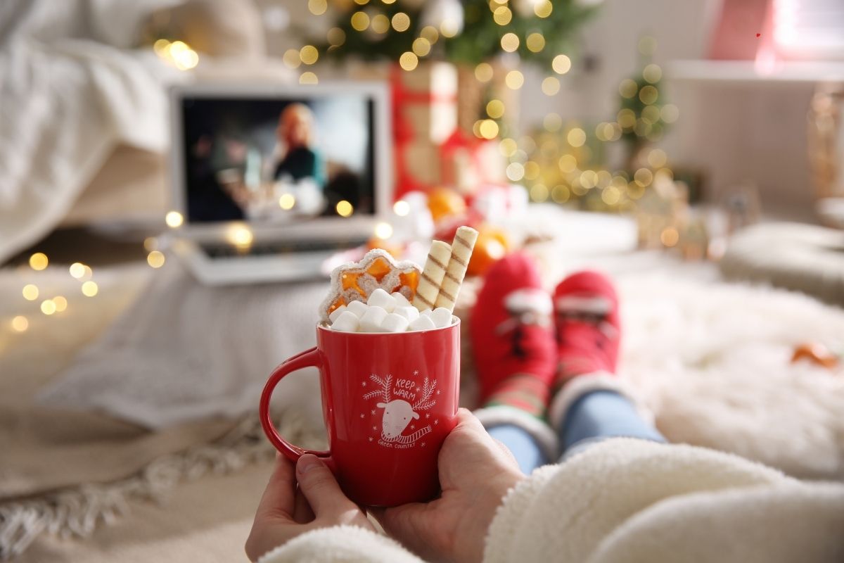 person holding hot chocolate in a room decorated for Christmas with Christmas movie on laptop